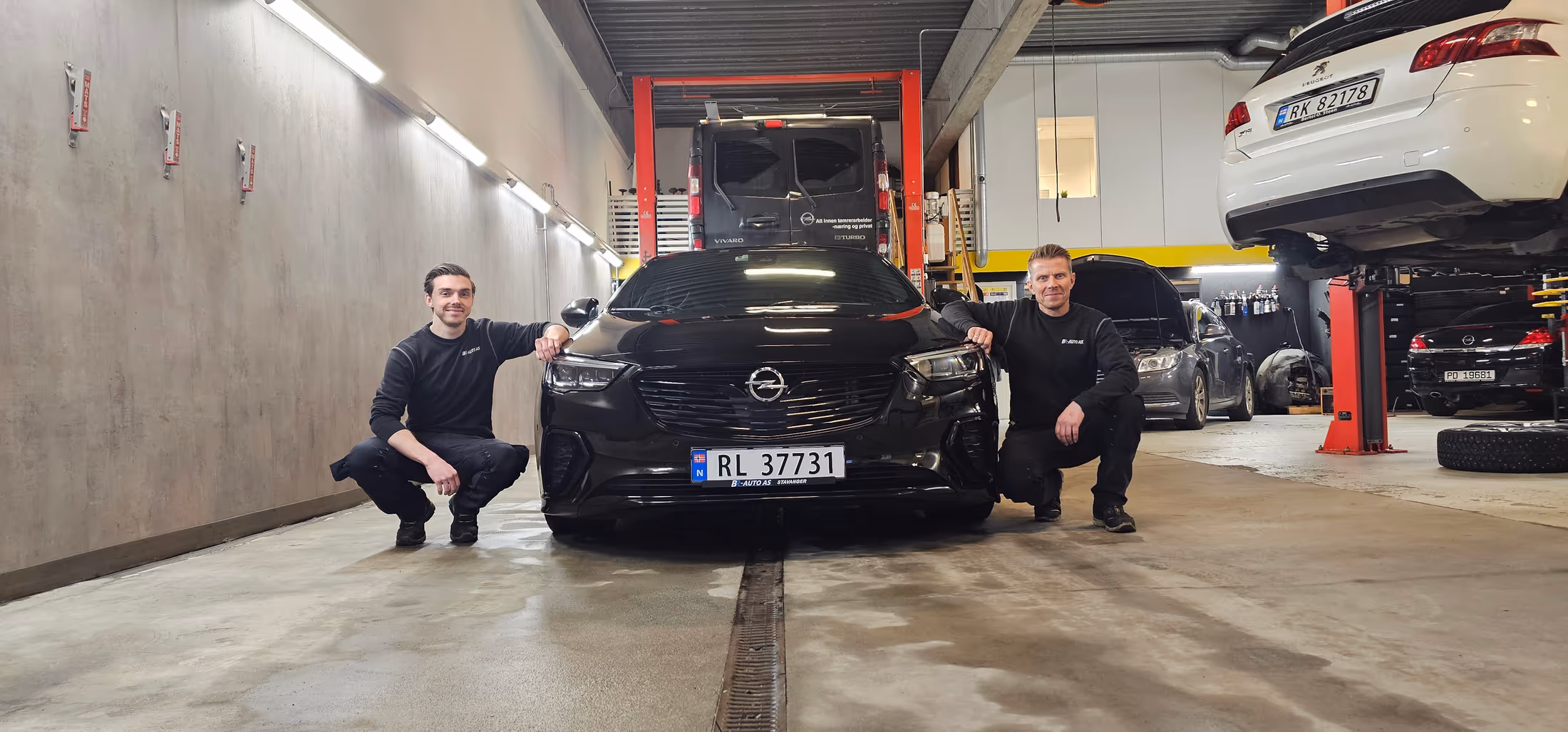 Einar og Mads Bru ved en svart Opel inne i verkstedet til BR-Auto på Mariero i Stavanger, med biler på løftebukk i bakgrunnen.